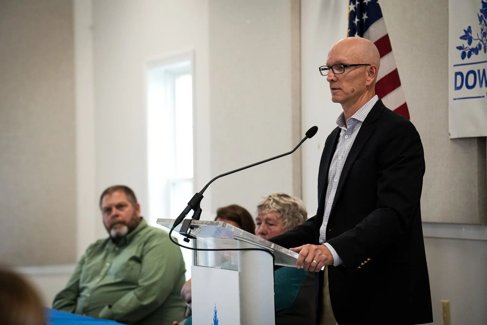 A man in glasses and a black blazer speaking at a clear podium with a microphone, with seated people and an American flag in the background.