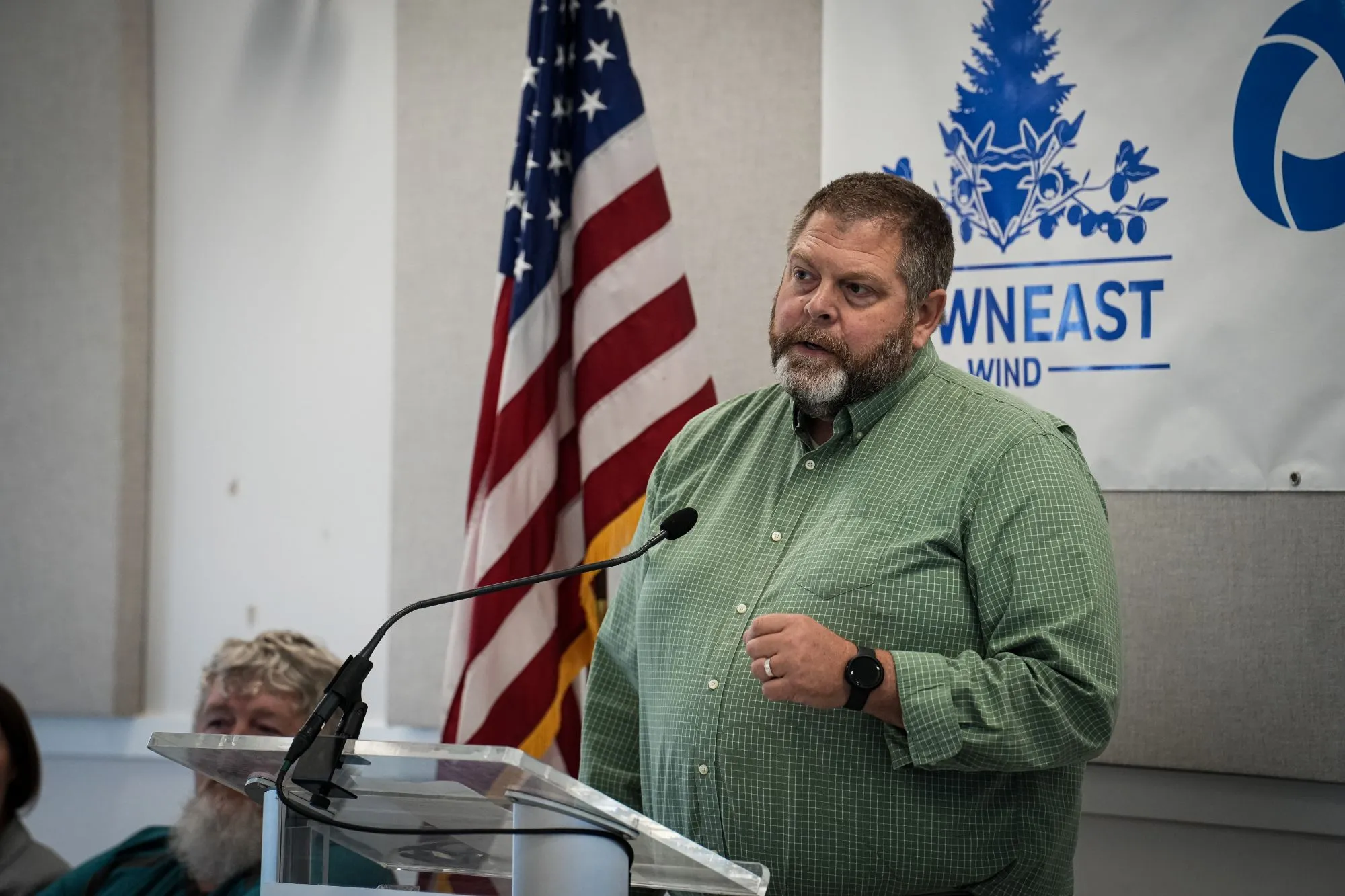 Bearded man in green shirt speaking at podium with American flag and Maineowneast Wind banner behind him.