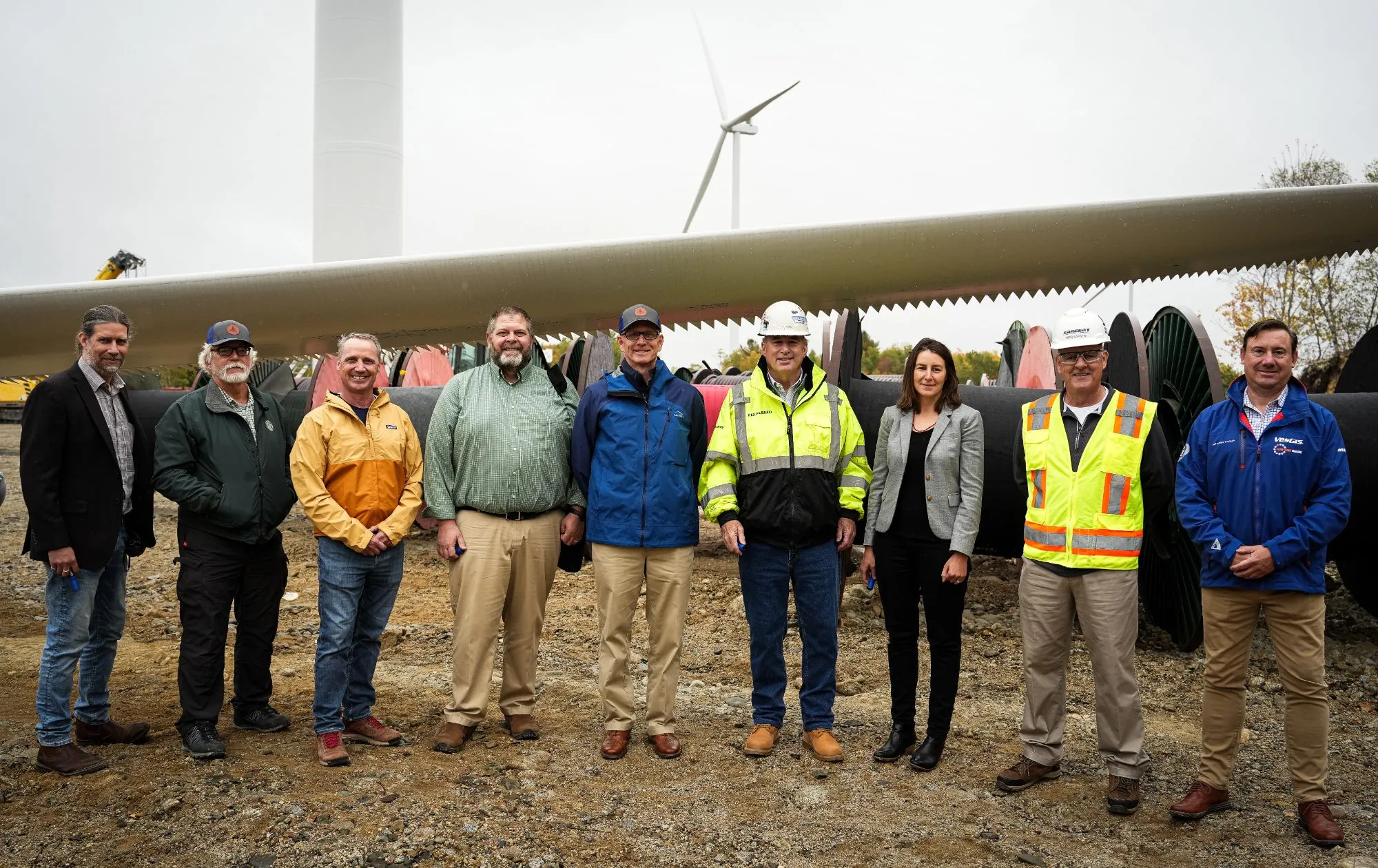 Group of nine people standing in front of a large wind turbine blade outdoors, with a wind turbine tower and another wind turbine in the background on a cloudy day.