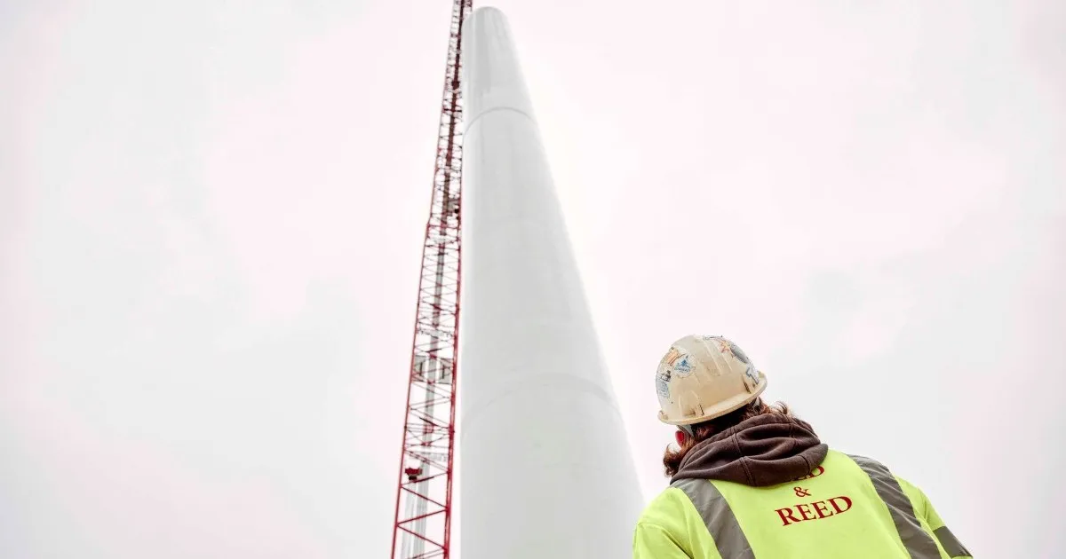 Construction worker in a yellow reflective jacket and hard hat looking up at a tall white wind turbine tower with a red crane beside it against a gray sky.
