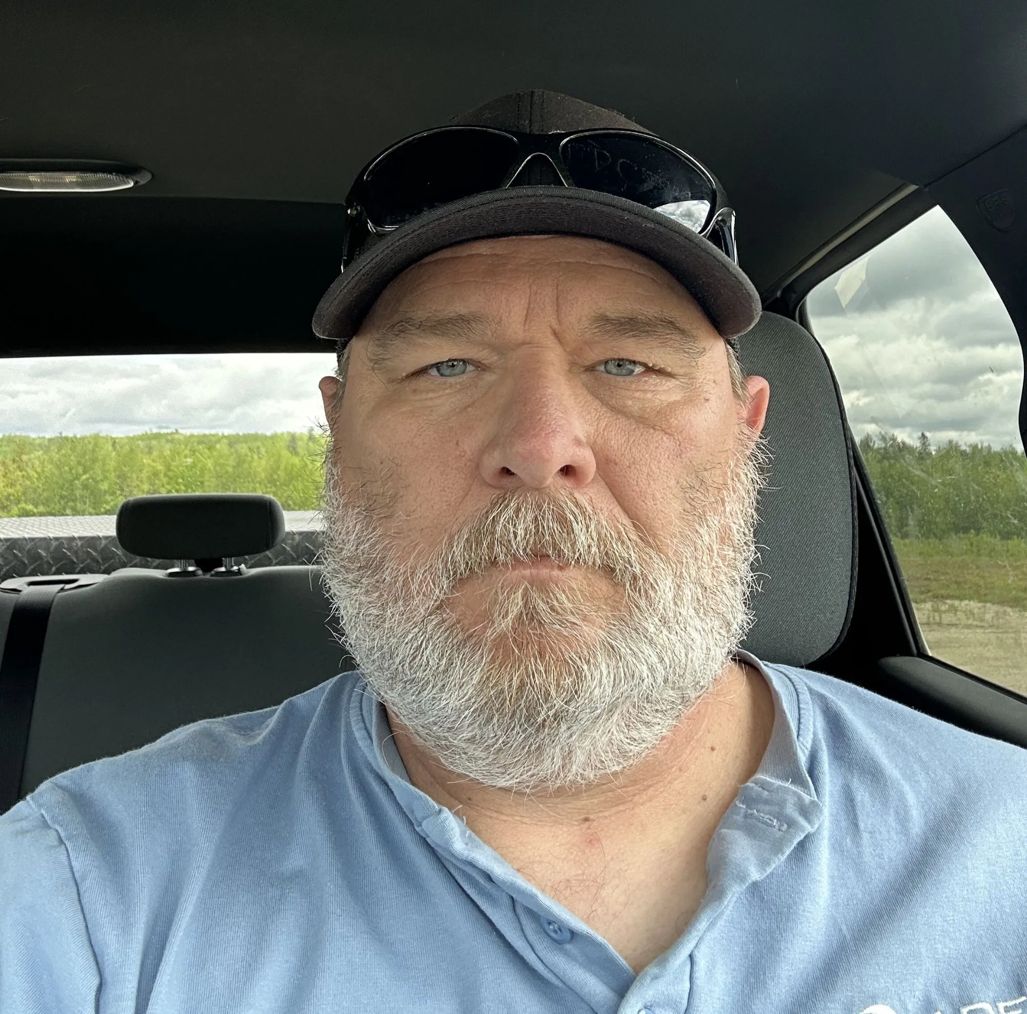 Middle-aged man with a white beard and mustache wearing a black baseball cap with sunglasses resting on top and a light blue henley shirt, sitting inside a car with a view of green trees and cloudy sky outside the window.
