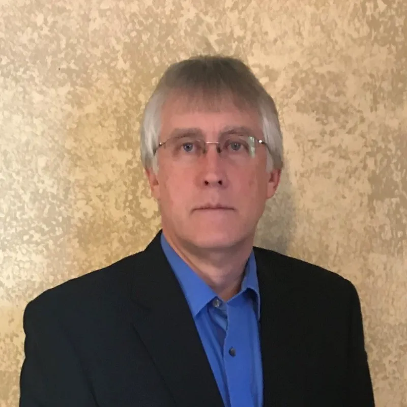 Portrait of a middle-aged man with gray hair and glasses wearing a black blazer over a blue collared shirt against a textured beige wall.