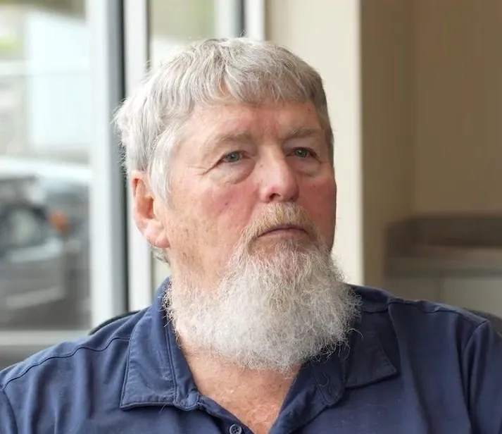 Older man with gray hair and a full white beard wearing a navy blue collared shirt, looking slightly to the side in a softly lit indoor setting.