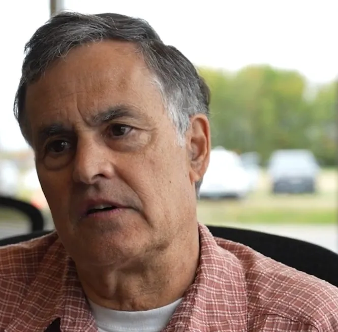 Middle-aged man with gray hair wearing a red plaid shirt and white undershirt, sitting indoors with blurred outdoor background.