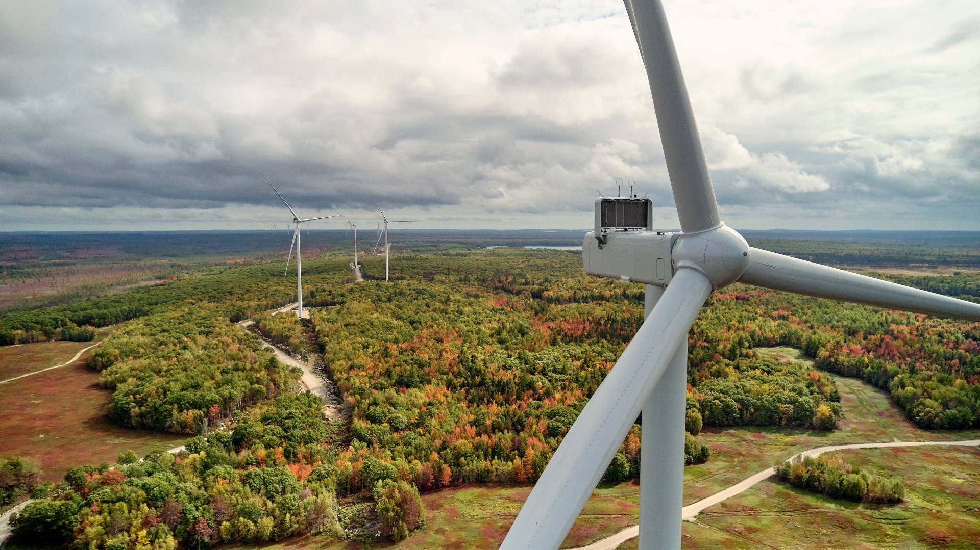 Aerial view of several large wind turbines on a forested landscape with fall foliage under a cloudy sky.