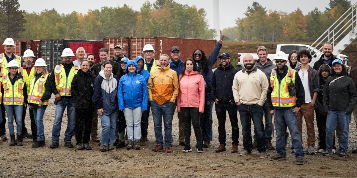 A group of diverse people standing outdoors on a dirt ground in front of stacked shipping containers and trees with autumn foliage; some wear construction safety vests and hard hats, others wear casual jackets and raincoats, and one person raises an arm in a cheerful gesture.