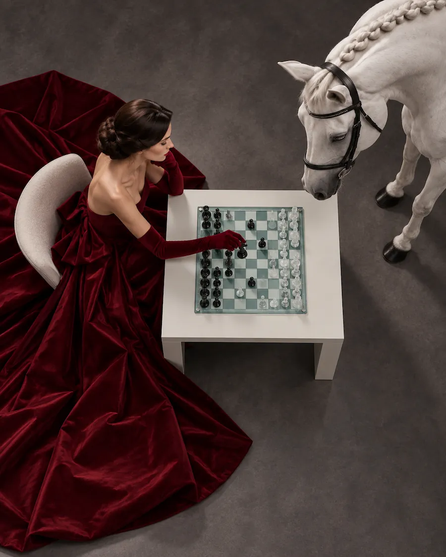 Overhead view of a woman playing chess as a white horse looks toward the board.