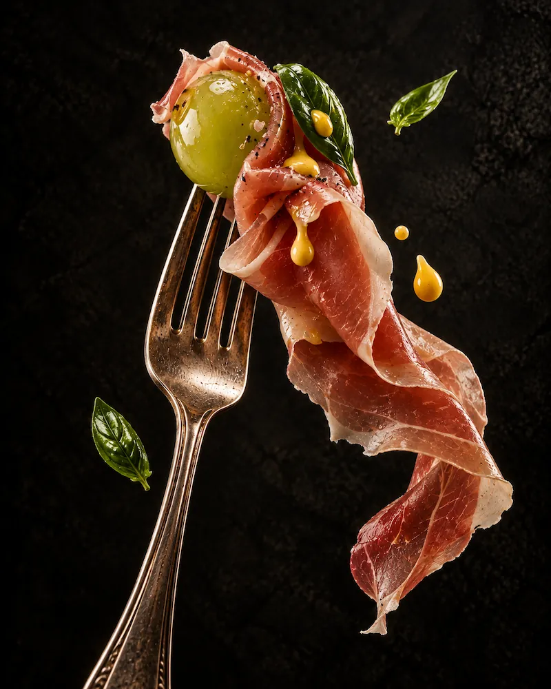 Prosciutto, a green olive and basil arranged around a fork against a dark background.