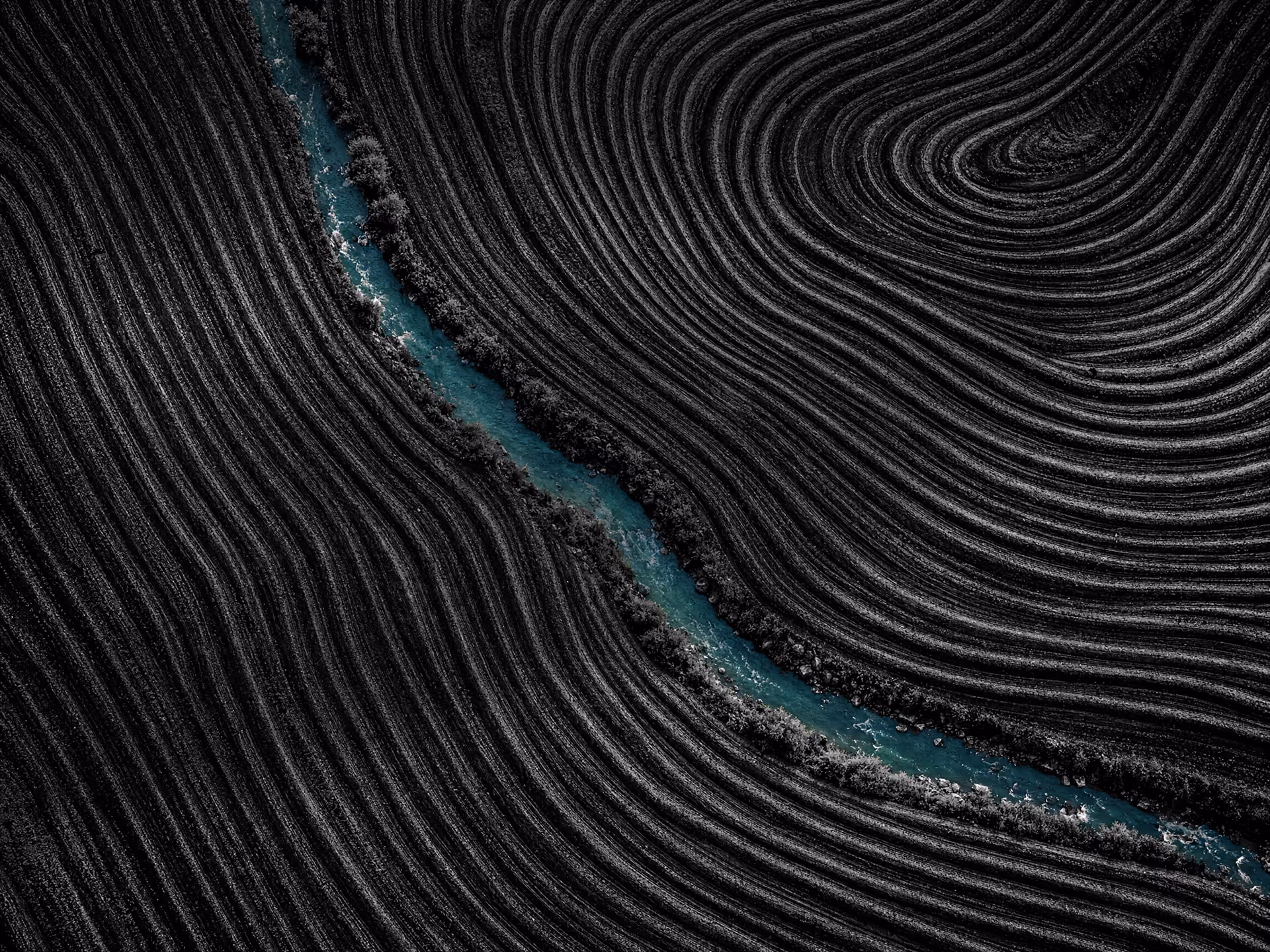 Aerial view of dark plowed agricultural fields with curving furrows, divided by a winding blue-green water stream bordered by shrubs.