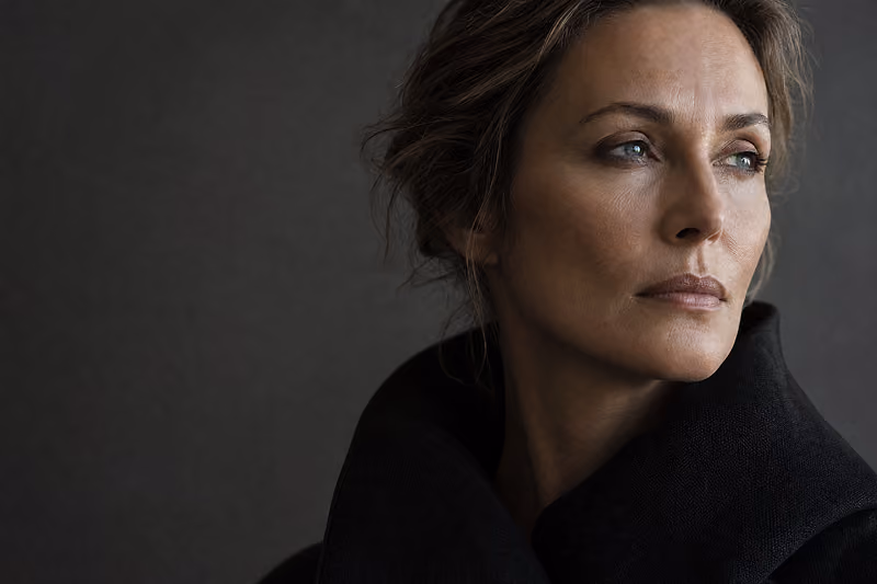 Portrait of a thoughtful woman with light skin and brown hair gazing to the right against a dark gray background, wearing a black high-collar garment.