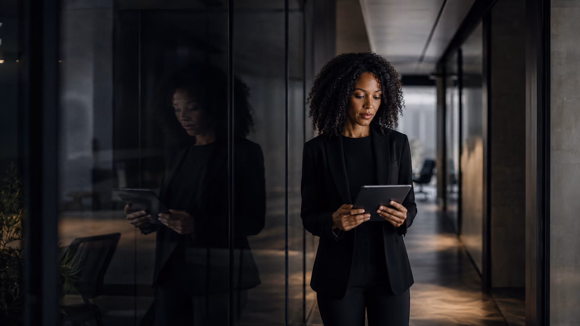 Woman with curly hair dressed in black business attire standing in a modern dimly lit office corridor, looking at and holding a tablet, her reflection visible on the glass wall beside her.