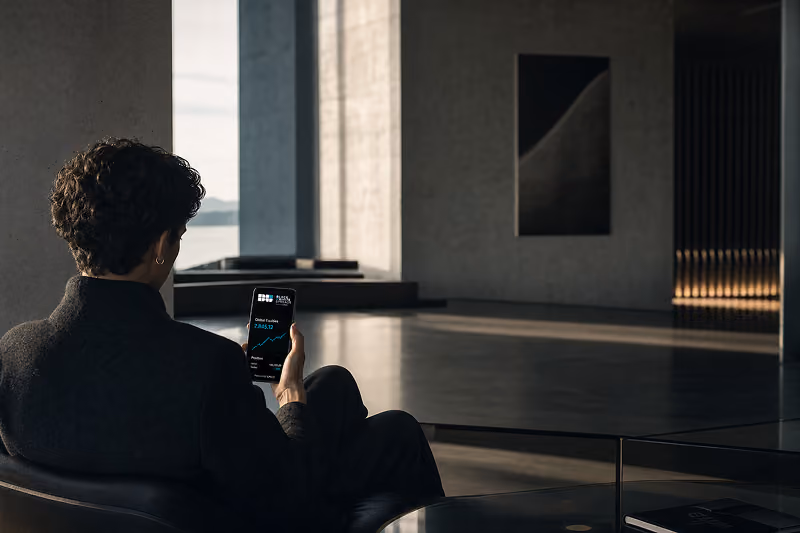 Person with curly hair wearing a dark coat sitting indoors and looking at a smartphone displaying a financial app with a stock price and graph visible.