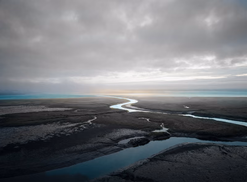 A winding river flowing through a dark, barren landscape under a cloudy sky with a faint glow near the horizon.