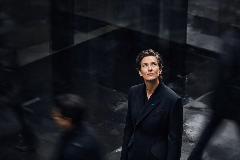 A woman with short hair wearing a dark blazer looks upwards in a dimly lit space with dark reflective surfaces, while blurred figures move around her.