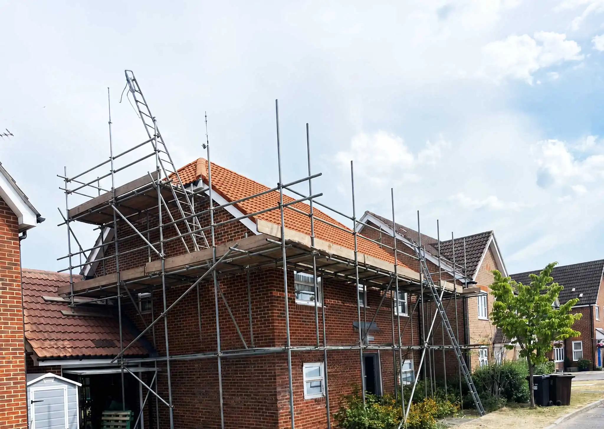 Completed residential roofing project with scaffolding, showing newly installed orange tile roof on brick house in suburban street setting