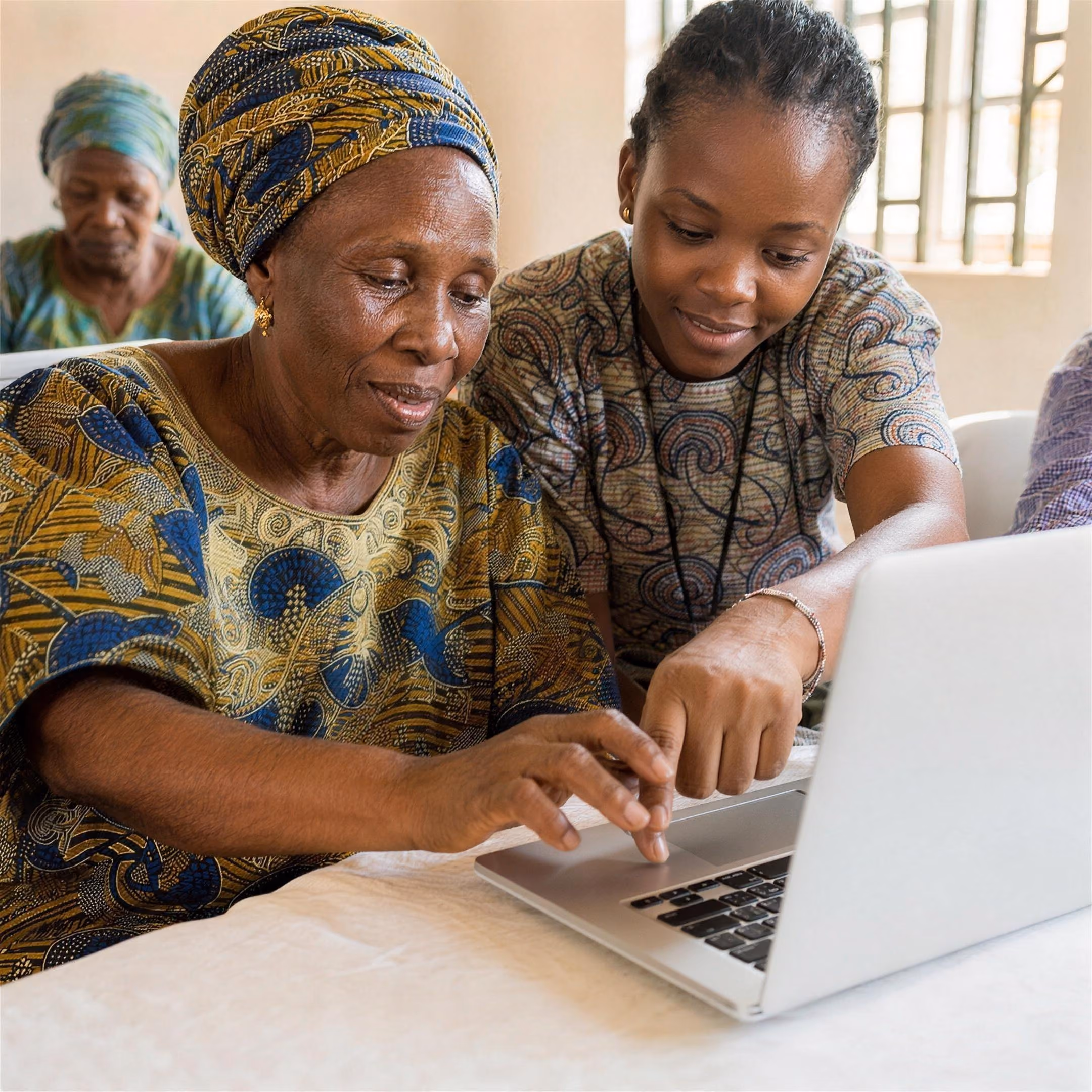 An older woman's hands rest on a laptop keyboard while a facilitator gently guides her hand to the trackpad.