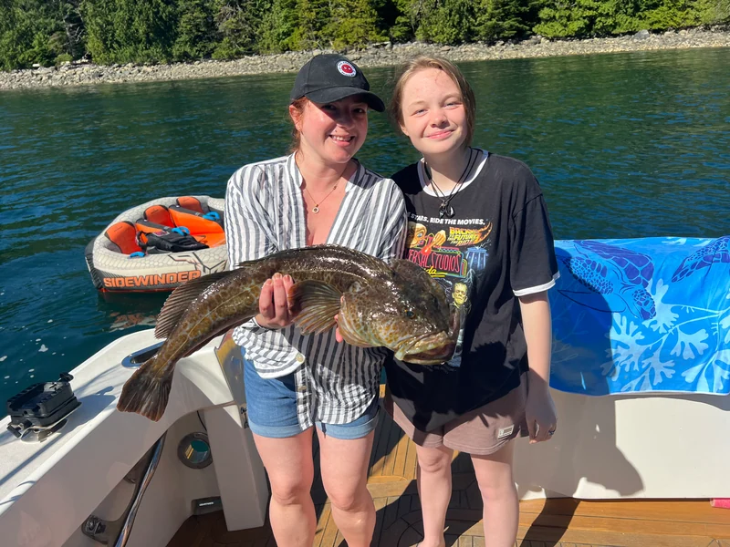 Two people holding a fish aboard a smaller boat in a wooded anchorage.