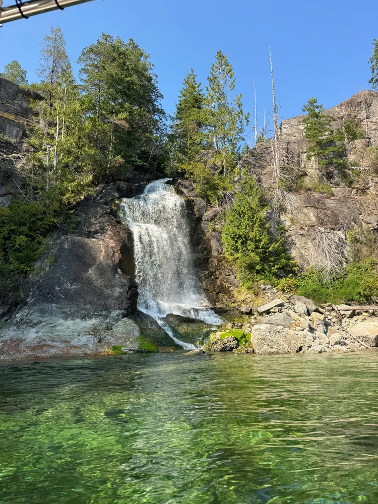 A waterfall drops into clear green water beside a wooded rocky shoreline.