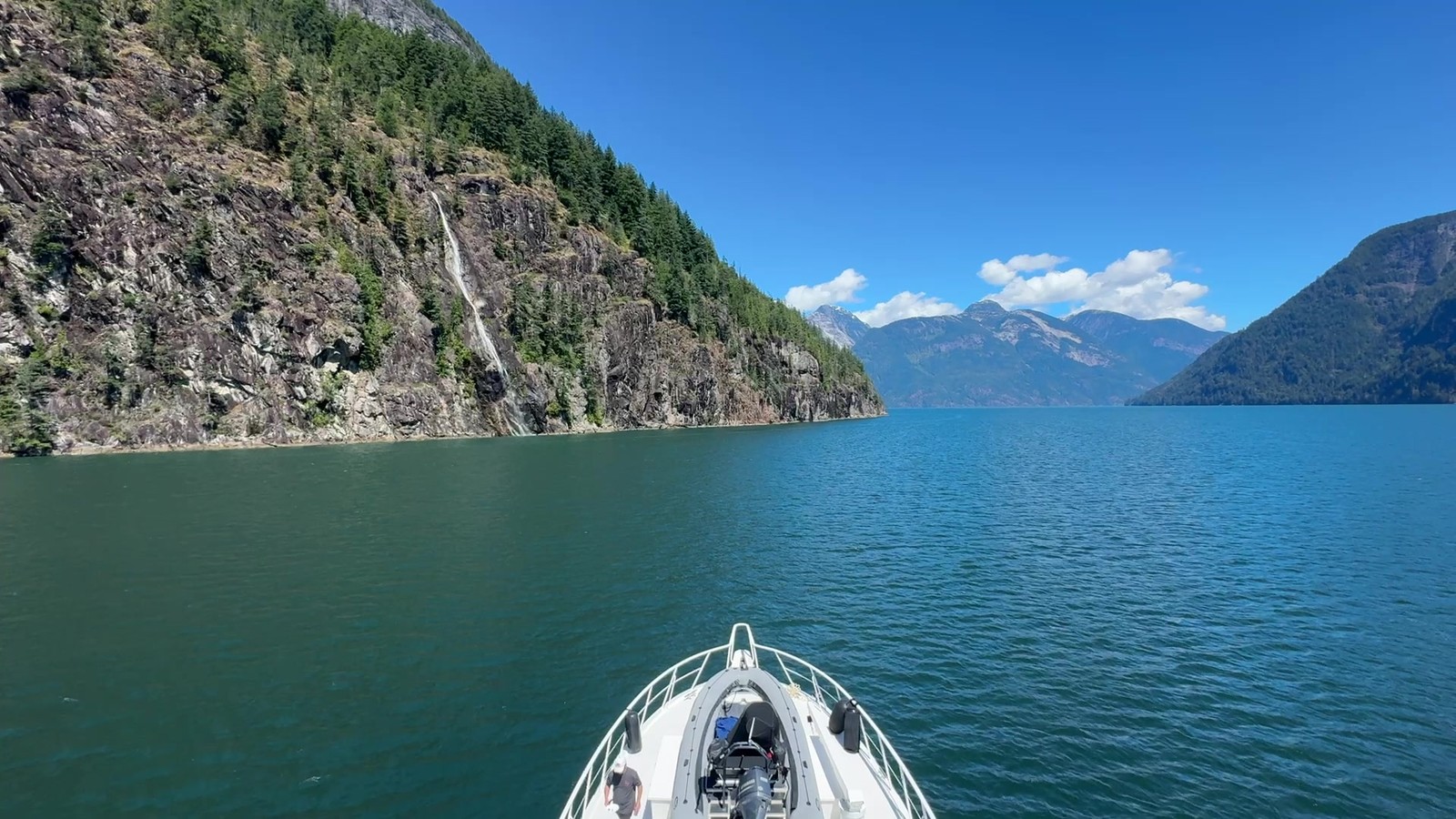 A waterfall beside a wooded cliff, seen over Surfrider's bow with mountains beyond