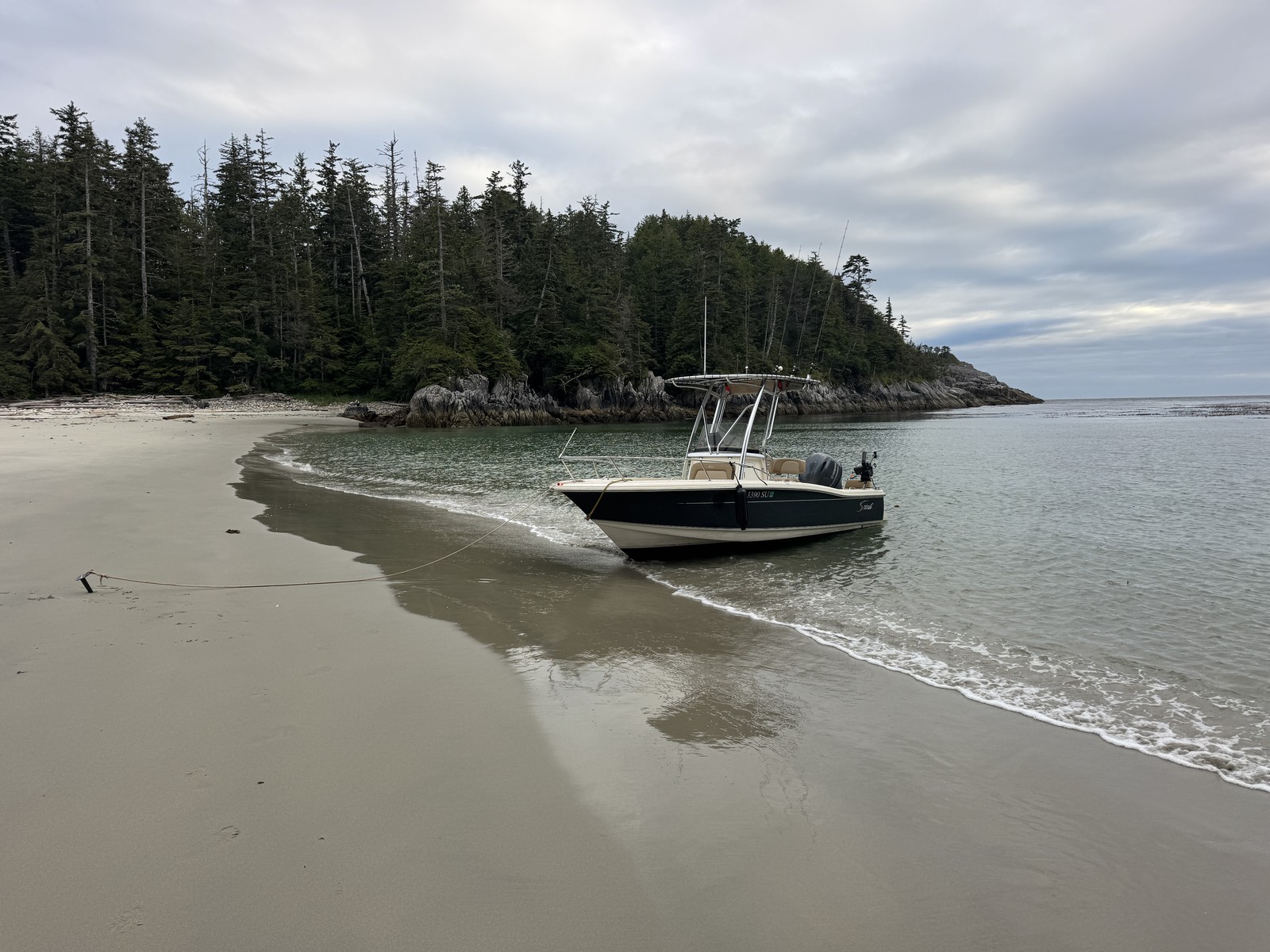 A center-console boat beside a pale-sand beach and wooded rocky shoreline at Hakai