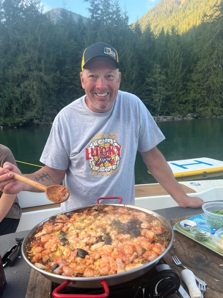 A smiling cook holding a spoon beside a large pan of paella aboard the yacht.