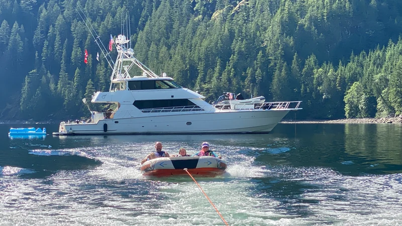 People riding a towable in front of Surfrider at a wooded anchorage.