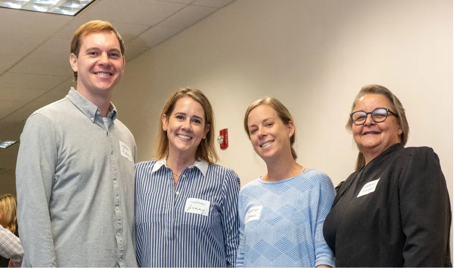 Four smiling people standing in a row indoors against a beige wall, wearing name tags, including one man in a gray shirt and three women in various tops.