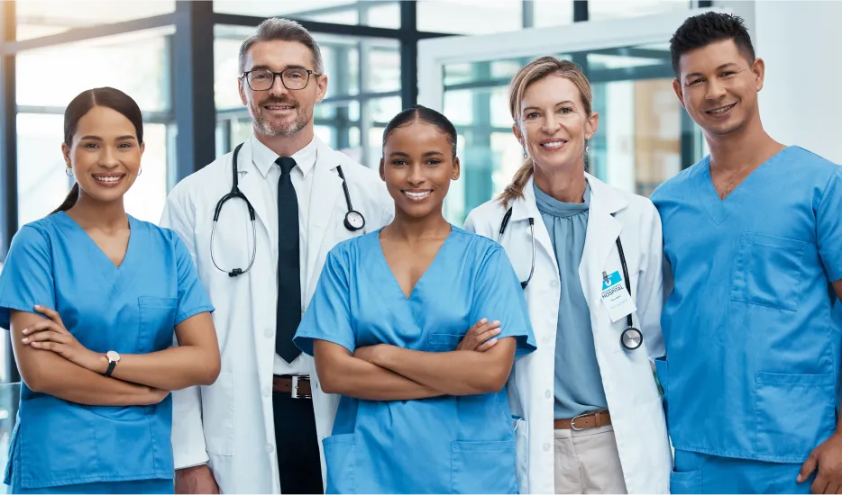 Group of five diverse healthcare professionals, three wearing blue scrubs and two in white coats with stethoscopes, standing in a bright hospital corridor smiling at the camera.