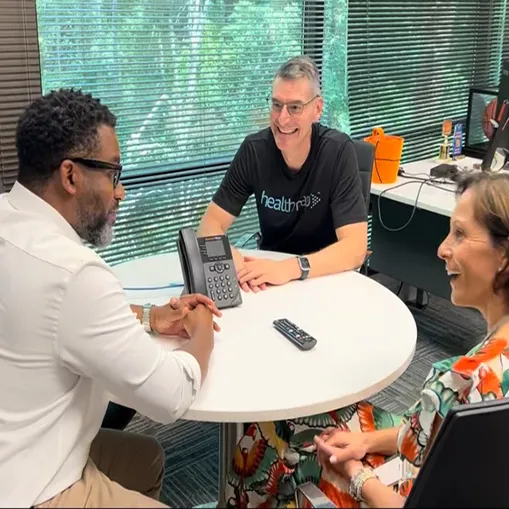 Three people sitting around a white round table in an office with large windows; a man in a black healthpad t-shirt smiles while talking, another man with glasses and a white shirt listens, and a woman in a floral dress smiles.