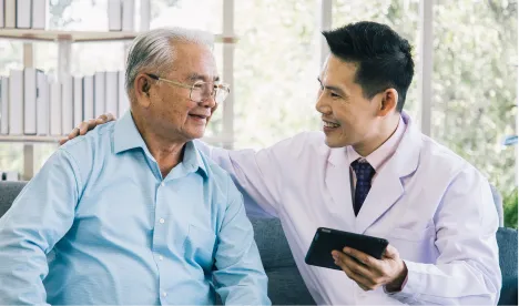 A doctor in a white coat holding a tablet and smiling while talking to an elderly man wearing glasses and a light blue shirt, sitting together in a bright room with large windows.