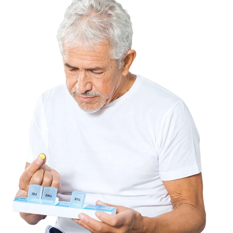 Older man with gray hair in a white t-shirt holding a weekly pill organizer and examining a yellow pill.