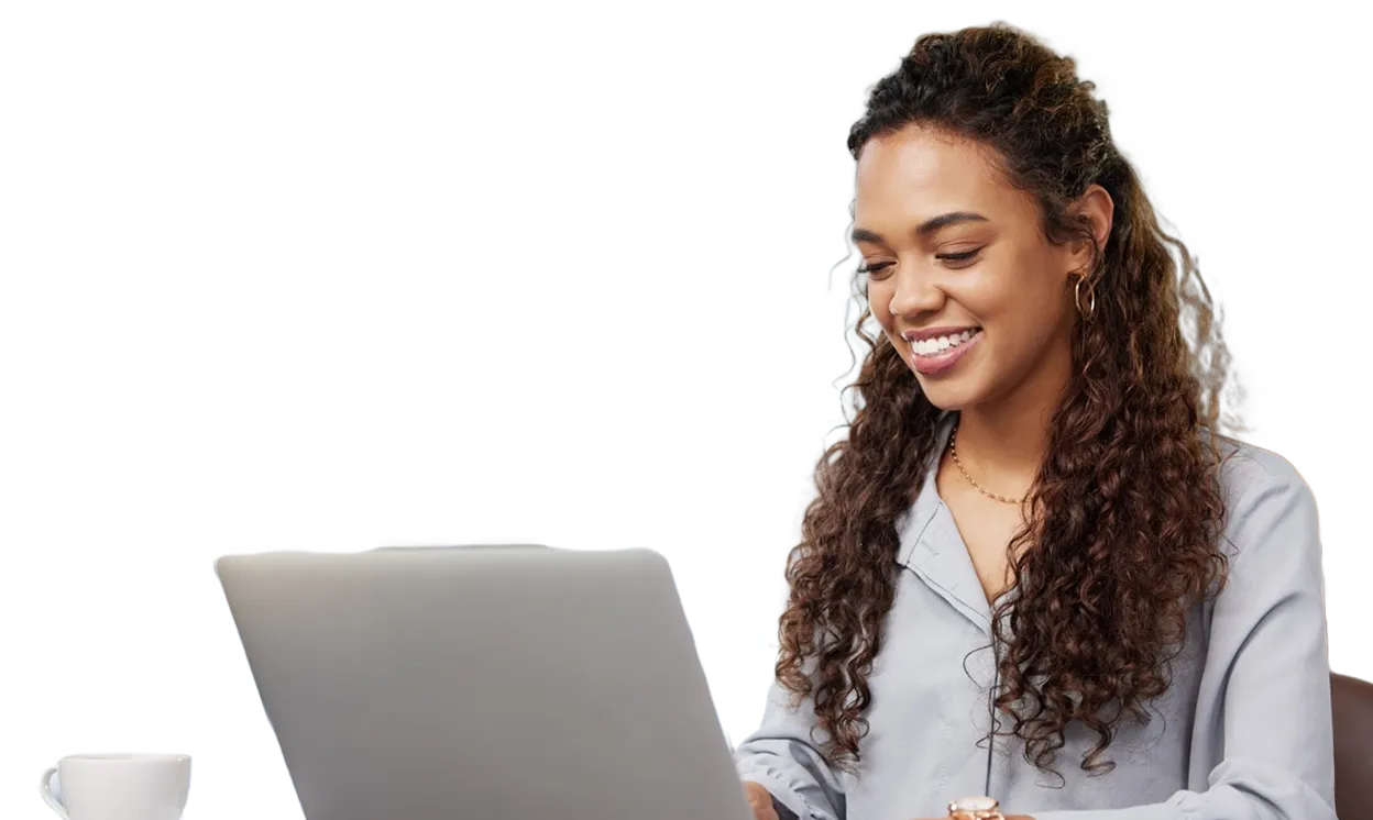 Smiling woman with curly hair working on a laptop, wearing a light gray top, with a white coffee cup nearby.
