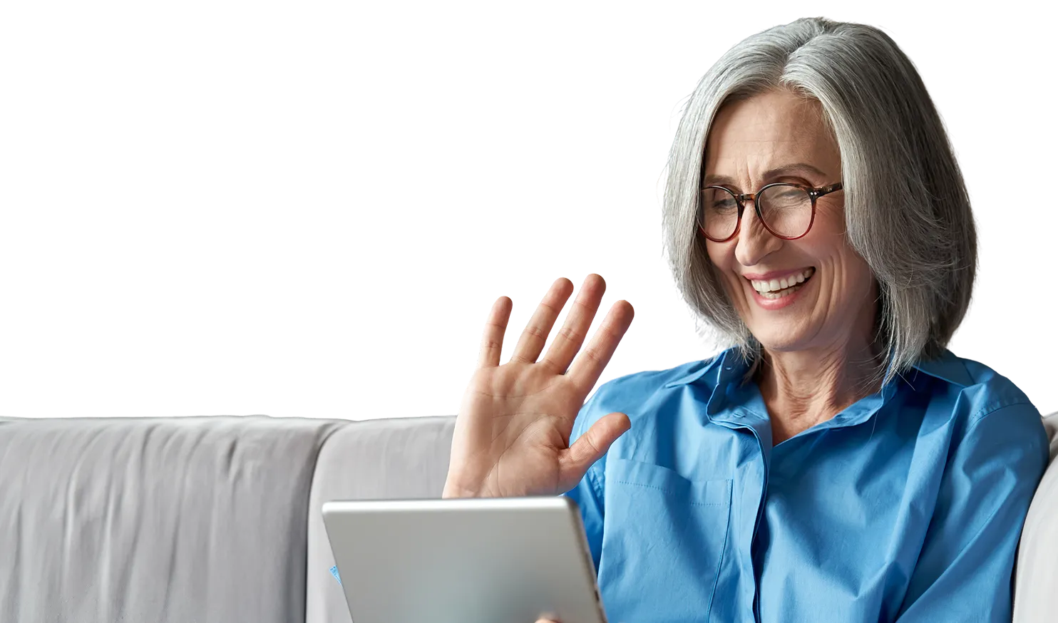 Smiling older woman with gray hair and glasses sitting on a light gray couch, waving while looking at a tablet.