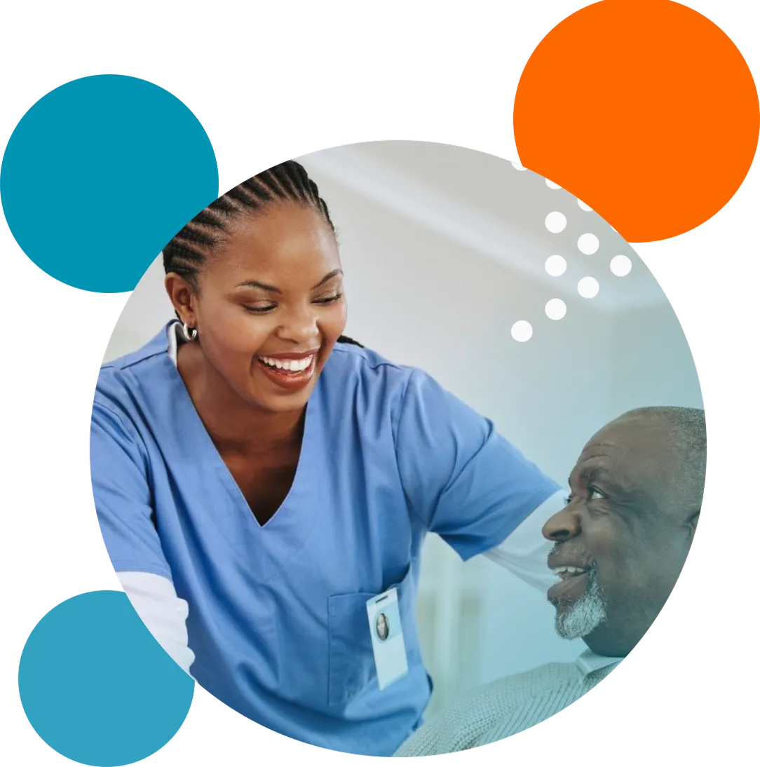 Smiling female nurse in blue scrubs with a name badge leaning toward and engaging with an elderly male patient who is looking up at her with a smile.