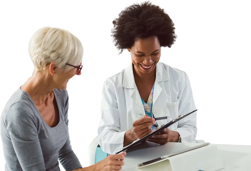 Smiling female doctor in white coat reviewing a clipboard and writing while speaking with an older woman with short white hair and red glasses.