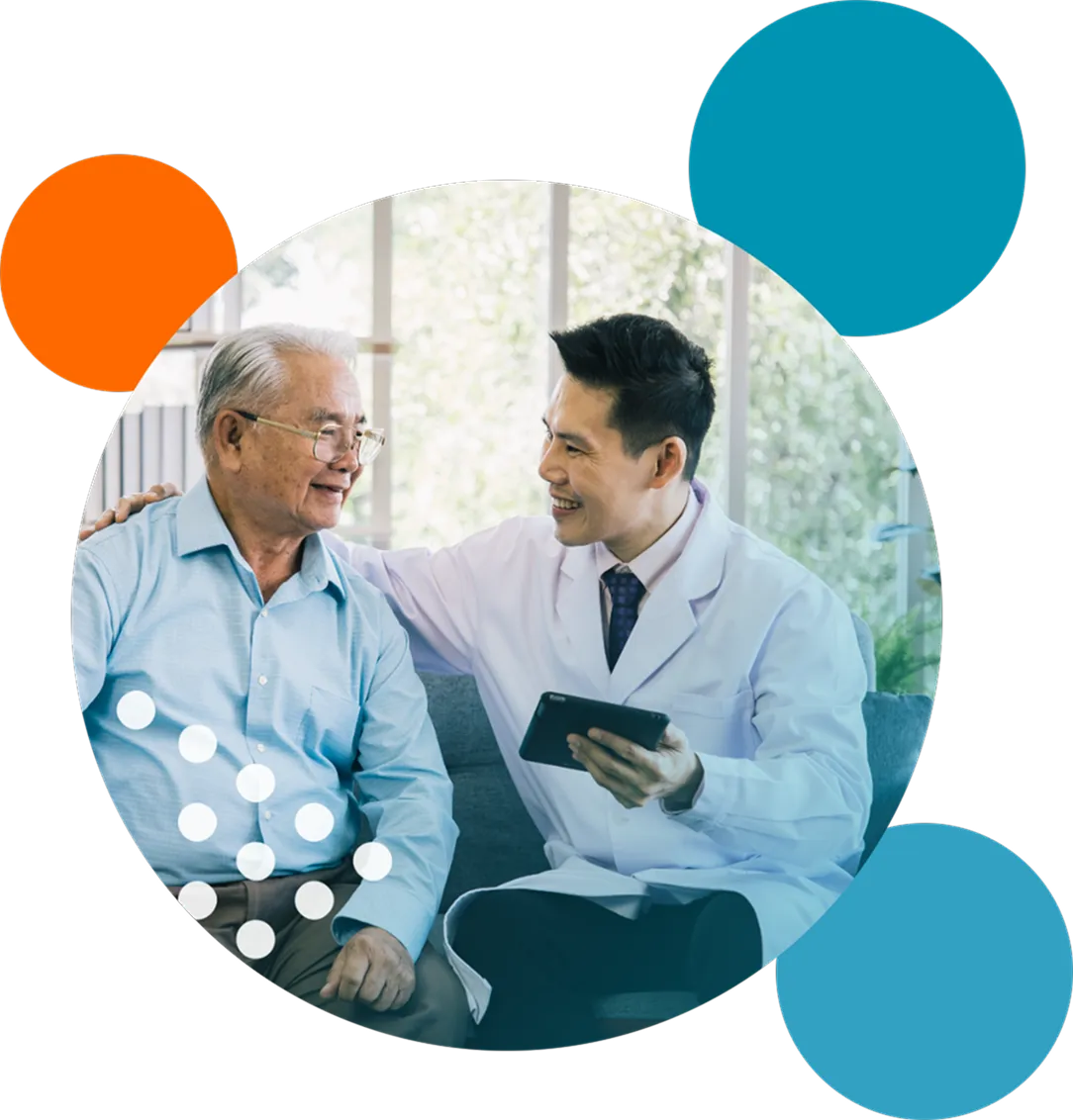 Smiling male doctor in a white coat sitting and holding a tablet with his hand on the shoulder of a senior man wearing glasses and a light blue shirt, having a friendly conversation in a bright room with large windows.