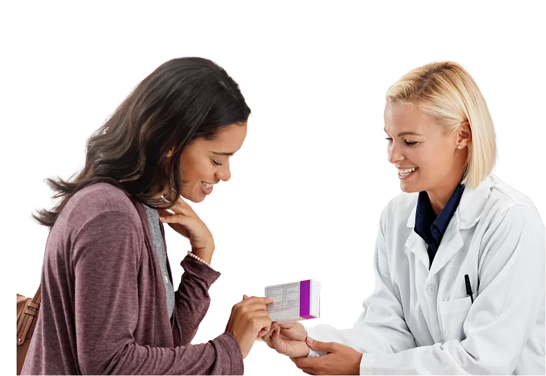 A smiling female doctor in a white coat shows and explains a medication box to a happy woman with dark hair wearing a purple sweater.