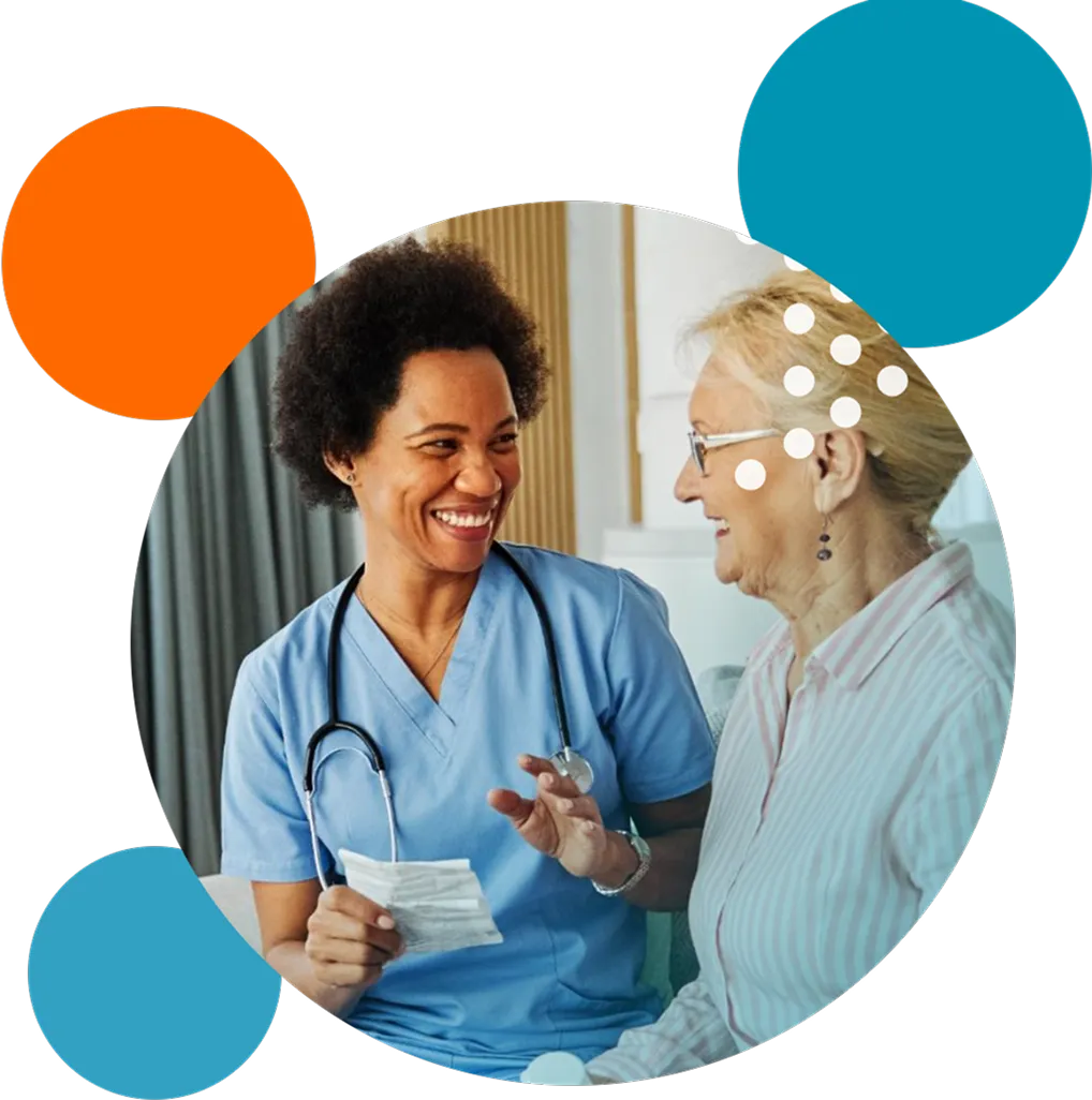 Smiling female nurse in blue scrubs holding a piece of paper while talking to an elderly woman wearing glasses and a striped shirt.