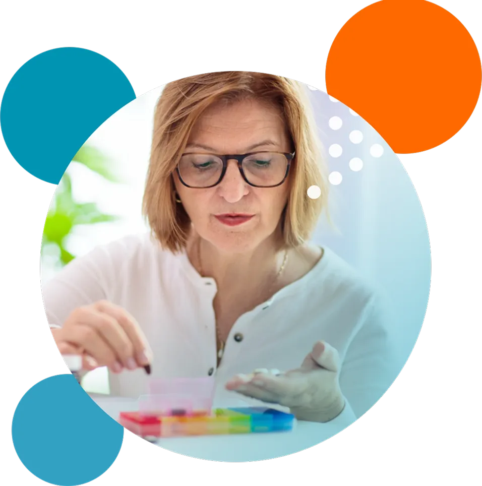 A middle-aged woman with glasses wearing a white shirt is organizing pills into a colorful weekly pill organizer.