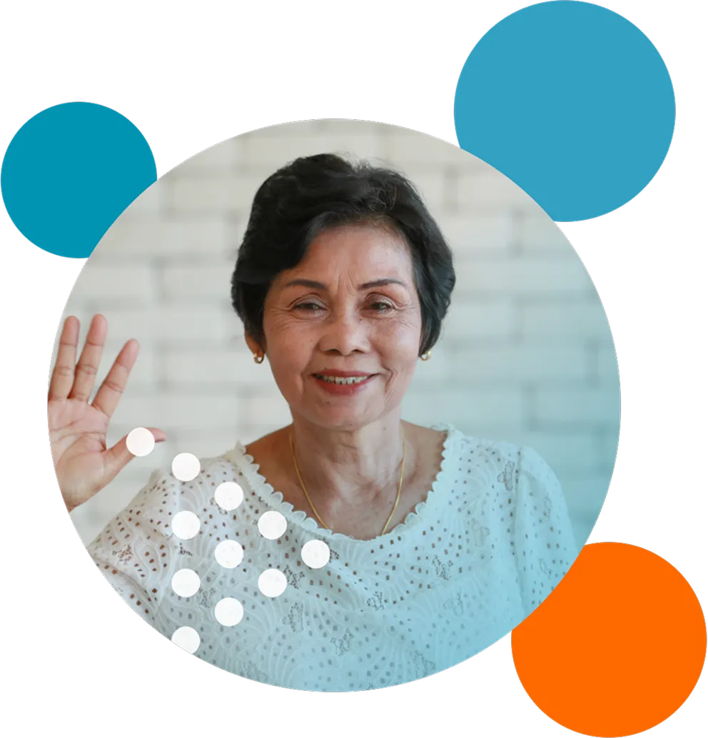 Smiling elderly woman with short dark hair wearing a white eyelet blouse and gold necklace, raising her right hand in greeting against a blurred light brick wall background with decorative blue and orange circles.