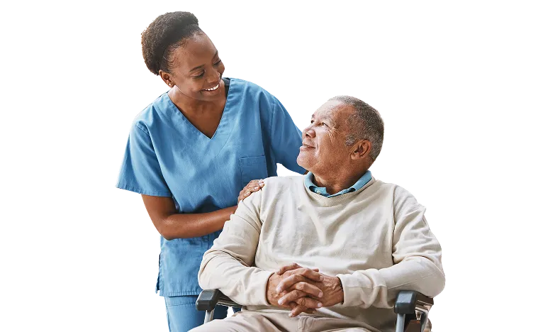 Smiling female healthcare worker in blue scrubs stands beside an elderly man in a wheelchair, looking at him warmly while resting her hand on his shoulder.