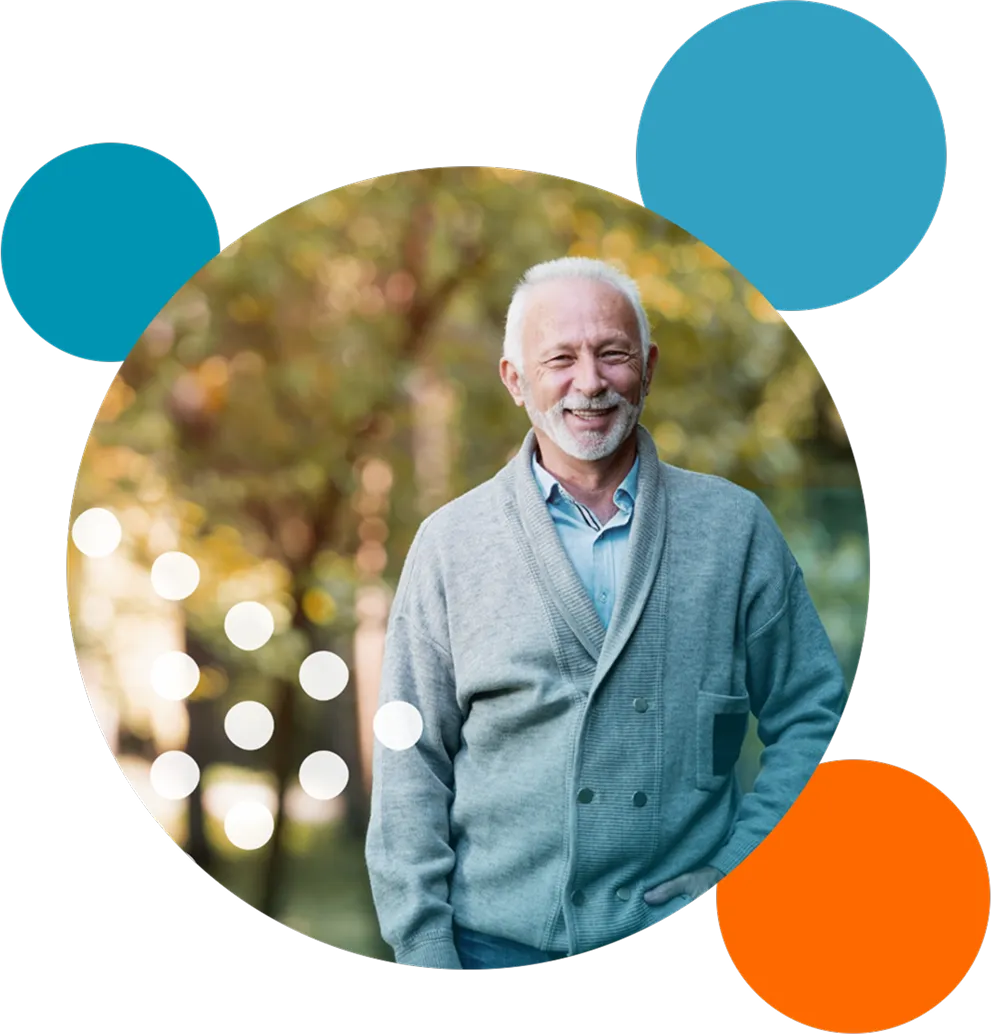 Smiling elderly man with white hair and beard wearing a gray cardigan and blue shirt standing outdoors with blurred autumn foliage in the background.