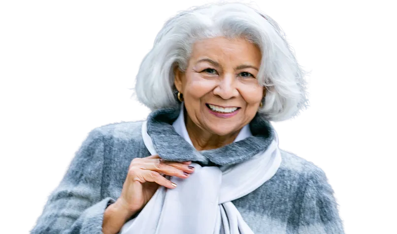 Smiling elderly woman with white hair wearing a grey coat and light scarf against a white background.