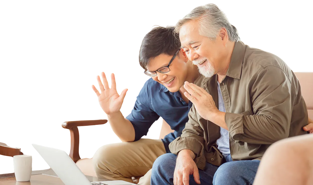 An elderly man and a younger man sitting on a couch smiling and waving at a laptop screen, appearing to have a video call.