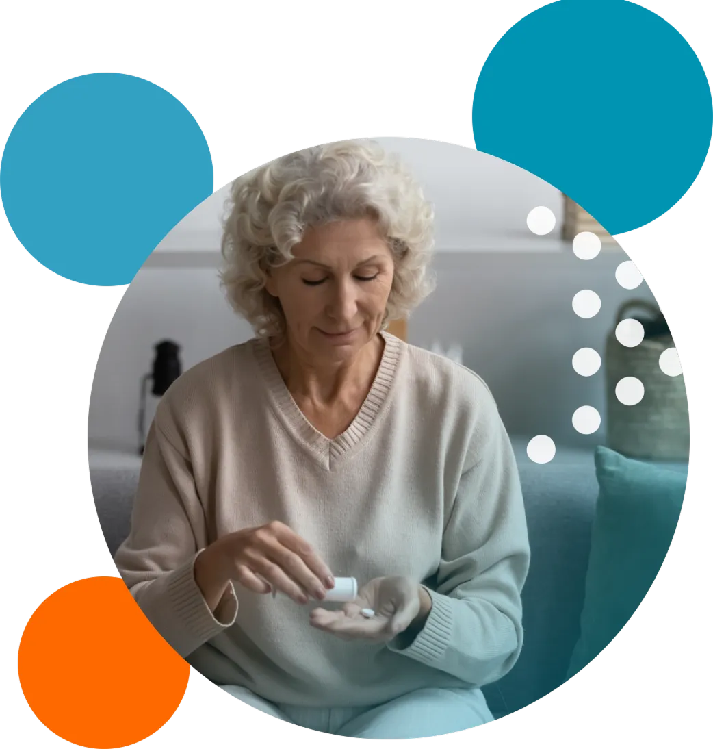Older woman with curly gray hair sitting on a couch, pouring pills from a white bottle into her palm, wearing a beige sweater.
