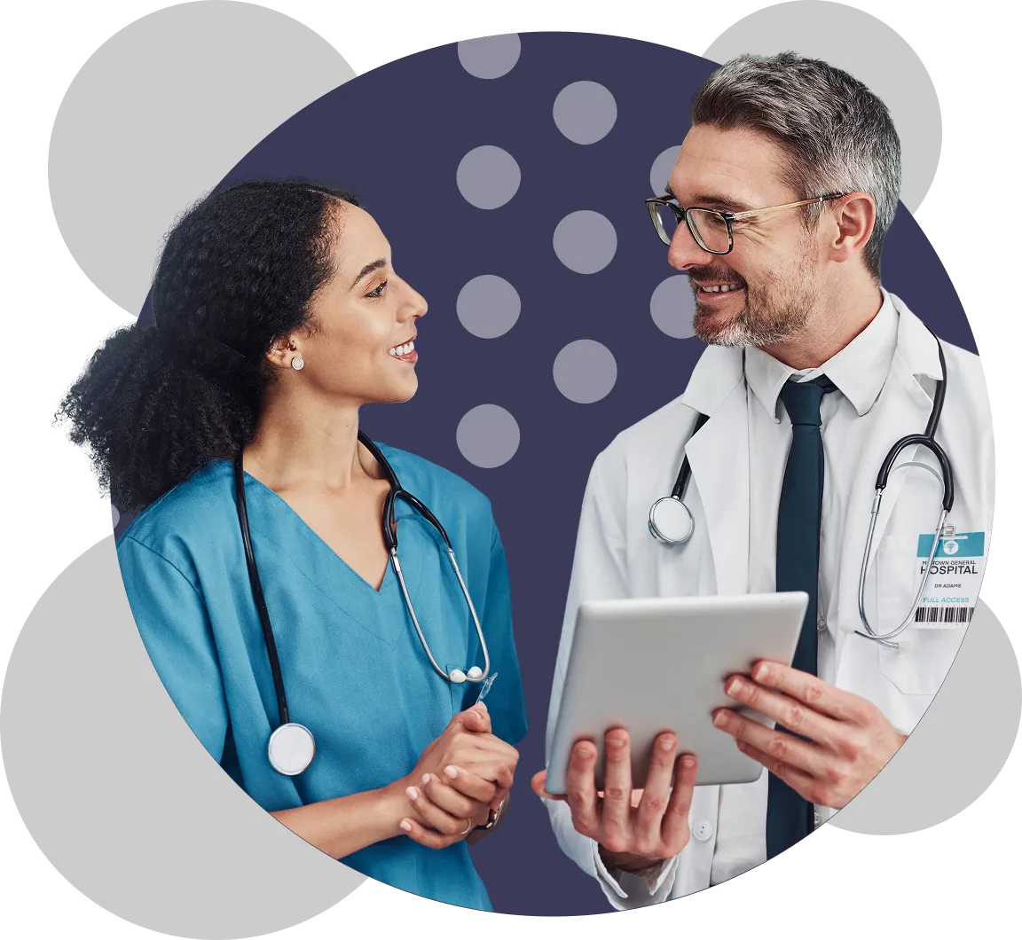 A female nurse in blue scrubs and a male doctor in a white coat, both wearing stethoscopes, smiling and discussing while the doctor holds a tablet.