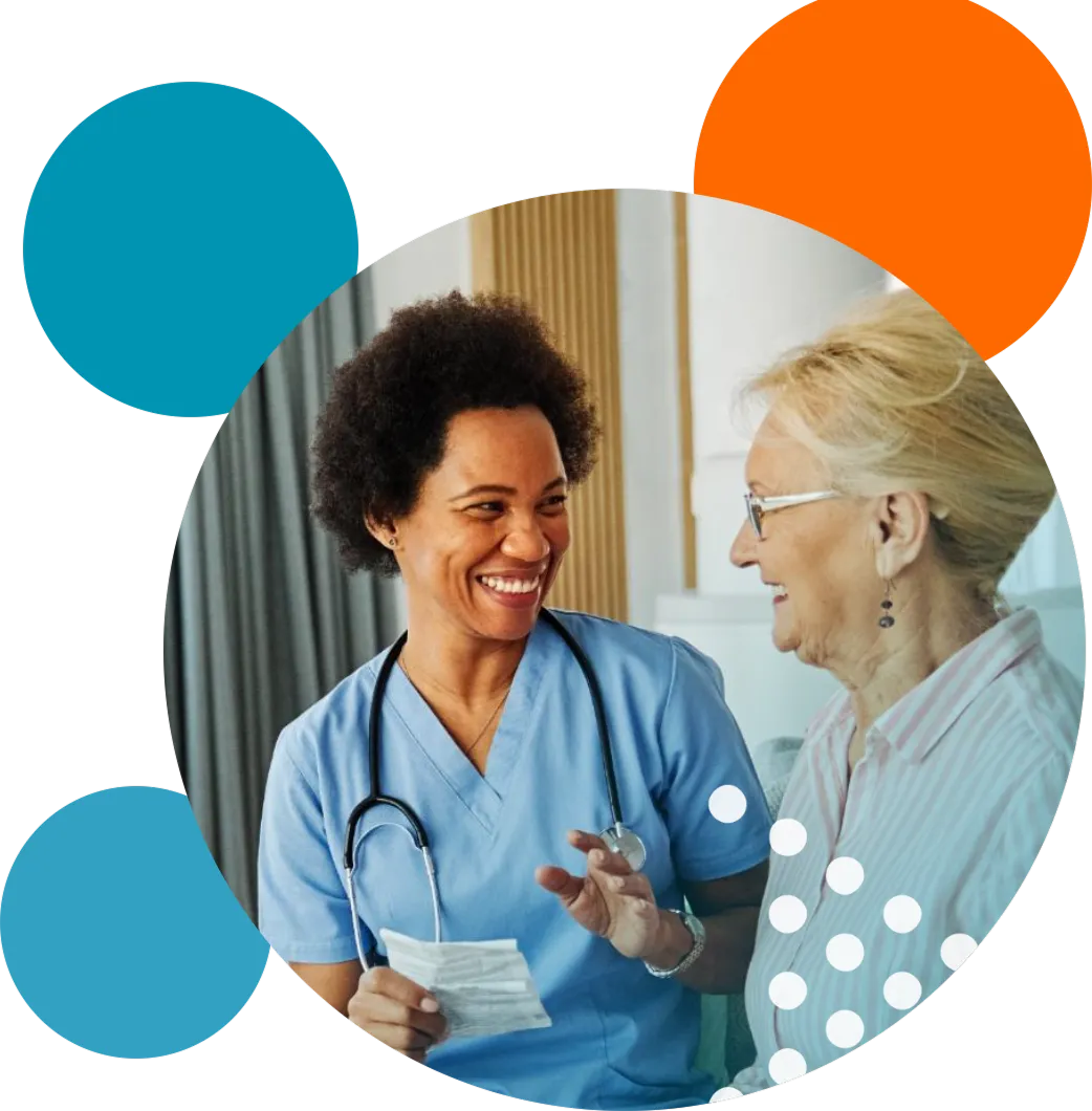 Smiling healthcare professional in blue scrubs with stethoscope talking to an elderly woman wearing glasses and a striped shirt in a bright room.