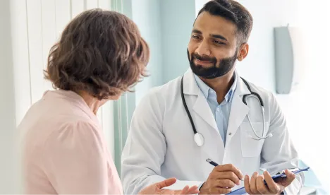 Male doctor with a beard and stethoscope listens and writes notes while talking to a female patient in a medical office.