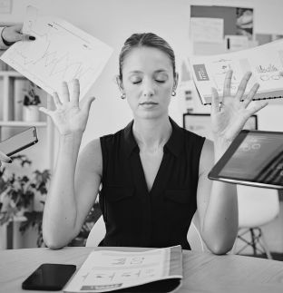 A woman sitting at a desk with eyes closed and hands raised in a calming gesture, surrounded by floating papers and a tablet, depicting stress or overwhelm in a work environment.