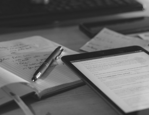 Black and white photo showing an open notebook with handwritten notes and a pen resting on it, next to a tablet displaying text, all on a wooden surface with a keyboard and papers blurred in the background.